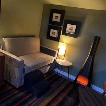 A cozy corner in a room featuring a grey armchair, a small table with a lamp, framed pictures on the wall, and a modern fireplace, showcasing interior design by Cypress Southern Homes in Tomball.